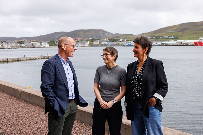 Three people in conversation outside with the sea and buildings visible behind them Three people in conversation outside with the sea and buildings visible behind them